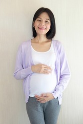 A pregnant Asian woman wearing casual clothes is standing smiling in a good mood.