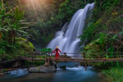 A young Asian tourist wearing a red dress stands on a bamboo bridge watching Pha Dok Siew Waterfall at Doi Inthanon National Park, Chiang Mai Province, Thailand.