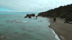 Top view of vacationing people on rocky seashore. Clip. Beautiful rocky coast with cliffs and reefs in blue water of sea. People walk along shore of reef sea on cloudy day