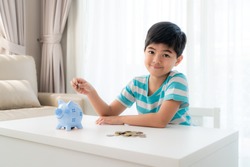 Little Asian boy insert a coin into blue piggy bank in white table at living room at home for the kid saving, investment money for his business future concept