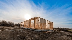 The structure of wood framing at construction site against a vast cloudy sky with bright sun behind. New build home at empty lot. framework ready for wall and roof install. Real estate development.