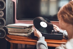 Young woman listening to music from vinyl record player. Retro and vintage music style. Girl holding analog record album sitting in room at home. Female enjoying music from old record collection