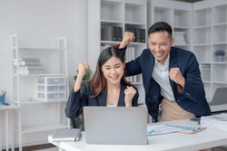 Two Asian business people looking at laptop with arms raised showing happy expression at office.