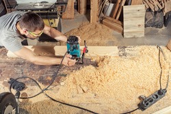 A young brunette man in safety glasses and a gray T-shirt is working a wooden board with a milling machine in the workshop, around a lot of wooden sawdust and wooden boards