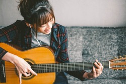 A beautiful asian woman smile while playing acoustic guitar and listening to music,sitting on sofa at home