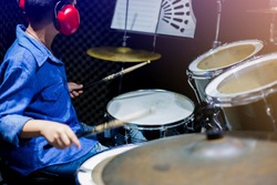 Asian boy put blue shirt and red headphone to learning and play drum set with wooden drumsticks in music room. The concept of musical instrument, Side view to drum player and selective focus.