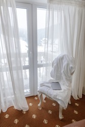 Hotel room in a mountain resort in winter with a white robe and a tablet on a chair, showing a nice bright view from the apartment through a window.