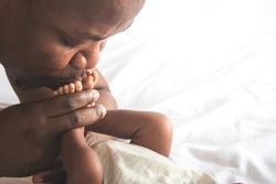 Close up images of An African American father kissing foot, his baby newborn, is laying on the white bed, with love and happy, concept to African American family and baby black skin newborn
