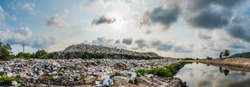 The panorama of large heap garbage dump extends parallel to the river,Garbage mountains with cloudy sky  back ground in day light,Waste has petroleum products are the major.