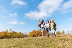 Three running girls at green grass at background of blue sky with clouds. Girlfriend concept. 3 young adult woman walk on summer field. Empty space for inscription.
