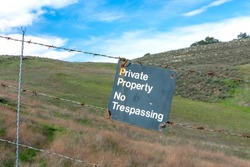 Private Property, No trespassing sign on barbed wire fence. Blurred green hills background under blue sky.