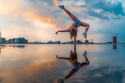 Female gymnast standing on one hand and keeping balance during dramatic sunset with reflection in the water of amazing clouds. Concept of Calisthenic, contortion and handstand 