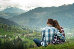 A couple in love sit in embraces on the top of mountain with a scenic view on the background. 
