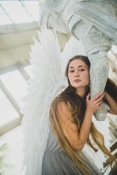 An angel girl with long hair and with large white wings behind her is holding a statue by her hand