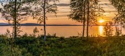 Scenic Panorama - Sunset over Umea river in mountains, summer sky with clouds highlighted by orange Sun. Blurry foreground with trees at coastline. Sunlight path on water. Storuman, Lapland, Sweden