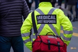 Close up of a medical first responder wearing a bright yellow coat with a grey reflective cross. The person is back on to the camera. There are people in the street in the background of the photo.