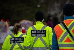 Medical first responders walking along a road wearing black wool stocking caps, yellow reflective coats with the medical first responder in grey letters and across. The EMT is carrying a first aid kit