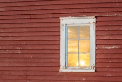 The exterior of an old red country wooden shed exterior wall with a white framed closed glass single hung window. In the window is a reflection of the cloudy sky with the orange-colored sun setting. 