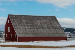 A large red vintage barn with multiple small closed glass windows with white trim. The sloping roof is made of old wood cedar shakes. The background is blue sky with clouds. There's snow on the field.