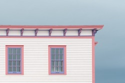 The roof section of a large white vintage wooden building with decorative pink and purple wood trim. There are three multi-pane windows on the top floor. The background is a dramatic blue cloudy sky.