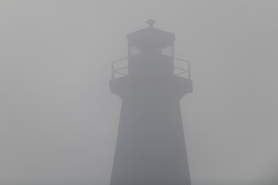 An abstract of an onshore circular style lighthouse. It's a stark white concrete tower with a green light beacon on top. The vintage building has a guardrail around the tower and is hidden by fog.