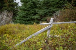 An old single grey and worn textured wood farm fence post with a broken rusty barb wire curled around the post with rust stains. There are multiple large rocks in the background among a grassy field.