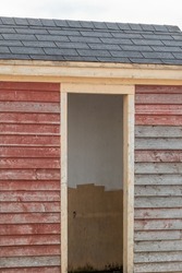 An open single doorway entrance to a small shed. The exterior wall is painted red with weathered boards, peeling paint and a grey shingled roof. The inside of the shed has white paint on the walls. 