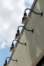 Multiple black colored barn lights in a row on the outside of a concrete building. The light fixtures have black shades with light directed downwards. There's a blue sky with clouds in the background.