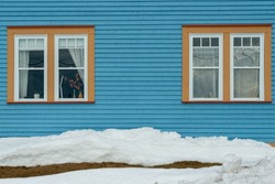 Two single hung windows with orange trim on a blue exterior wall of a vintage style building. The building has narrow clapboard on the wall and a mound of white snow on the ground in front. 