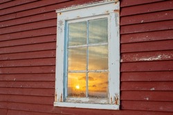 The exterior of an old red country wooden shed exterior wall with a white framed closed glass single hung window. In the window is a reflection of the cloudy sky with the orange-colored sun setting. 
