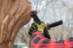 A male wearing a red and black plaid lumberjack coat with green and black gloves. He's using a wood carving power tool on an oak tree. The carving has lines and a line pattern. The tree has a bump.