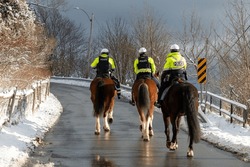 Three police officers of the mounted unit riding large horses on a street heading up a hill in winter. The cops are wearing bright yellow uniforms with police written across their backs and white hats