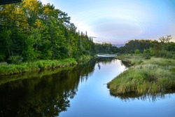 A scenic meandering river as the sun sets in the distance. The trees and lush green reeds are reflecting in the smooth water. The serene lake has lush riverbanks with green grass and a thick forest.