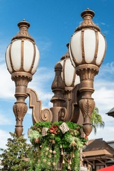 Three brass cast street lamps on a single decorative post of forged metal. The antique park light has a white textured milk glass. There's a decorative green Christmas garland hanging off the post.
