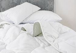 White pillows in cotton pillowcases lie on a rumpled bed. An airy white satin blanket lies casually on the bed. White new bedding