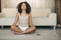 Young black woman practicing yoga at home, sitting in Easy Seat pose, her eyes closed, listens audio meditation by headphones with the phone