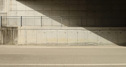 Concrete block wall of an underpass with a footpath and a railing in the middle. Side light and shadow. asphalt road in front. No people. Background for copy space.