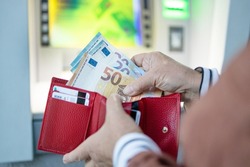 Unrecognizable female puts out a 50 and 20 euro banknote from a red leather wallet near the ATM on the street.
