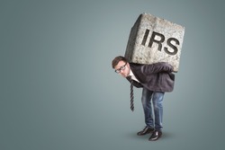 Businessman bending under a heavy stone with the letters IRS printed on it - tax office concept