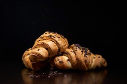 Three golden chocolate croissants and dark chocolate pieces on black background. Fresh and delicious French pastry, bakery concept, close up image. Copy space	