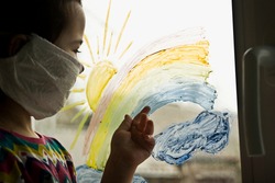 A small child draws a colored rainbow on a glass window. The kid at home on self-insolation during quarantine looks at the village through a painted rainbow on glass