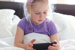 Little girl sitting up in bed using smart phone during the morning. 