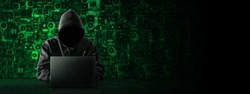 Hacker with a hidden face and with a laptop sits at the table, blue background. There is an hourglass on the hacker's desk. Place for text. High quality photo
