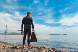 a diver in a wetsuit stands on the seashore. Diver before diving into the ocean, in the hands of a mask and fins for diving. High quality photo