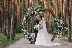 Stylish groom in a suit and a cute brunette bride in a white dress in the forest near a wedding wooden arch decorated with flowers. Wedding portrait of the newlyweds.