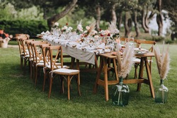 A large, long, decorated, wooden table and chairs, covered with a white tablecloth with dishes, flowers, candles, stands on the green grass in the park, in the forest in nature. Wedding banquet.