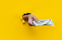 A right female hand appears in the hole in torn yellow paper and squeezes several hundred-dollar bills. Concept of poverty, benefits, scholarships, and stinginess. Isolated. Copy space.