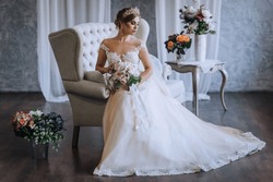 A beautiful blonde princess bride in a white lace dress with a crown, a diadem sits on a sofa in the studio, indoors with a bouquet in her hands. Wedding photography, close-up portrait.