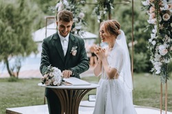 Stylish bearded bride and groom in a white dress are smiling while standing at the ceremony against the backdrop of a decorated arch. Wedding photography, portrait of the newlyweds.