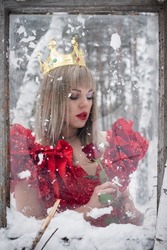 Young woman in the red dress with red rose flower in hand and golden crown looks through the frozen window. Ice queen concept.